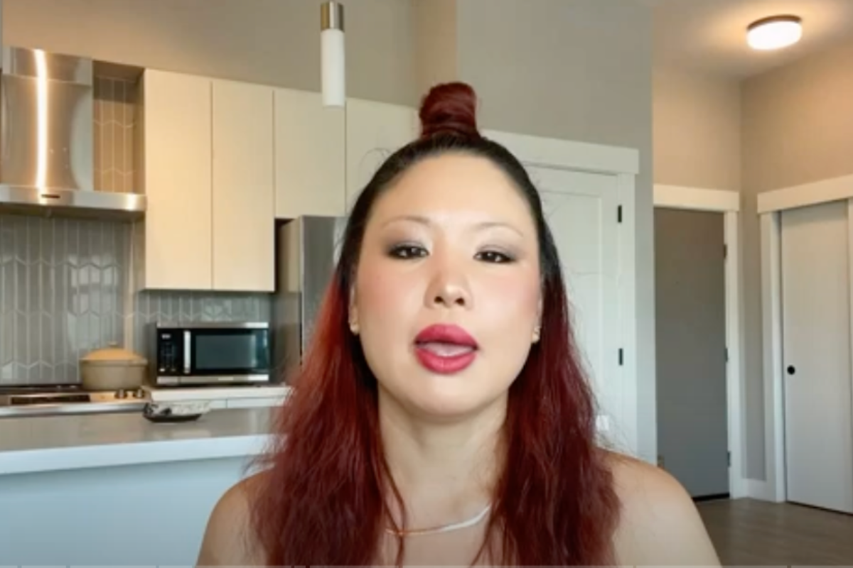 A woman with long red hair and bold makeup speaks while seated in a modern kitchen setting.